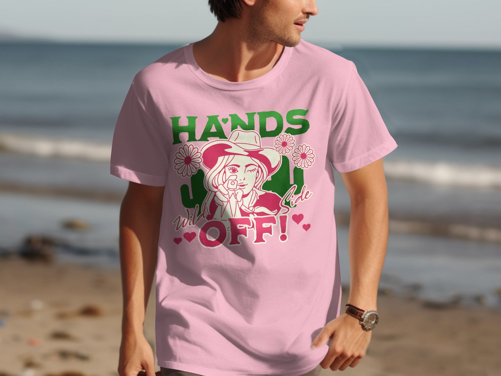 A man stands on a beach wearing a classic fit pink T-shirt by Garment Graphics. It showcases a cartoon woman in a hat with cacti and flowers, featuring HANDS OFF! prominently. Ocean waves accentuate the vibrant Hands Off T-Shirt design.