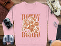 The Garment Graphics pink sweatshirt, ideal for rodeo lovers, showcases a cowgirl embodying cowboy spirit on horseback with Howdy Rodeo in bold brown text. Her hat says Not a Beginner. Surrounded by vintage items like a camera and gloves, it encapsulates the essence of Howdy Rodeo Sweatshirts.