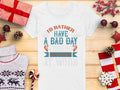 A Garment Graphics white fishing T-shirt with the words Id rather have a bad day fishing than a good day at work is displayed on wood. Its surrounded by gifts, pine cones, a sweater, an ornament, and berries. Available in womens sizes, its perfect for fishing fans.