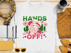 The Garment Graphics white T-shirt flaunts a unique design with a woman in a hat, stylized flowers, and bold HANDS OFF! Wild Side text, paired with a scarf, notebook, coffee cup, yellow handbag, pen, and sunglasses for the perfect look.