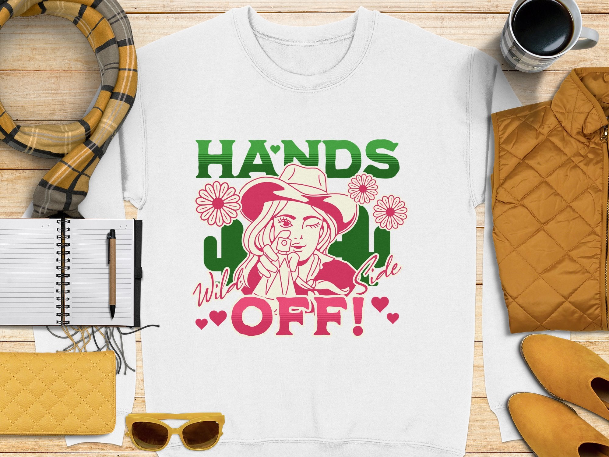 The Garment Graphics white T-shirt flaunts a unique design with a woman in a hat, stylized flowers, and bold HANDS OFF! Wild Side text, paired with a scarf, notebook, coffee cup, yellow handbag, pen, and sunglasses for the perfect look.