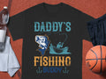 Garment Graphics black t-shirt showcases a vibrant fish and person in a boat graphic with Daddys Fishing Buddy in green and yellow, ideal for fishing fans. Nearby are a clear water bottle, white towel, black shorts, and an orange basketball on a reddish surface.