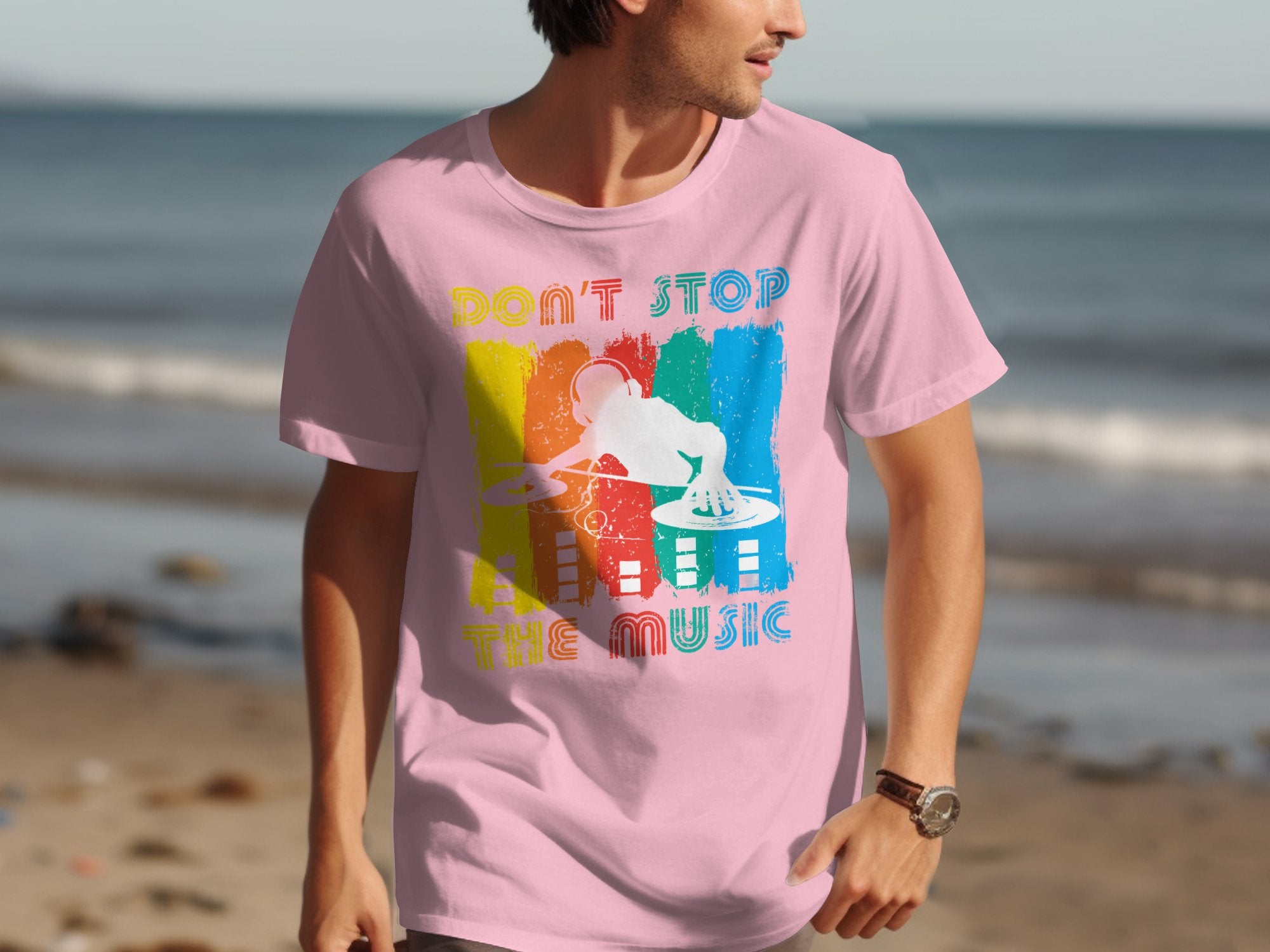 Wearing a Garment Graphics pink t-shirt with a vibrant DJ graphic and Dont Stop the Music, a man strolls along the beach, embodying the spirit of music lovers against ocean waves.