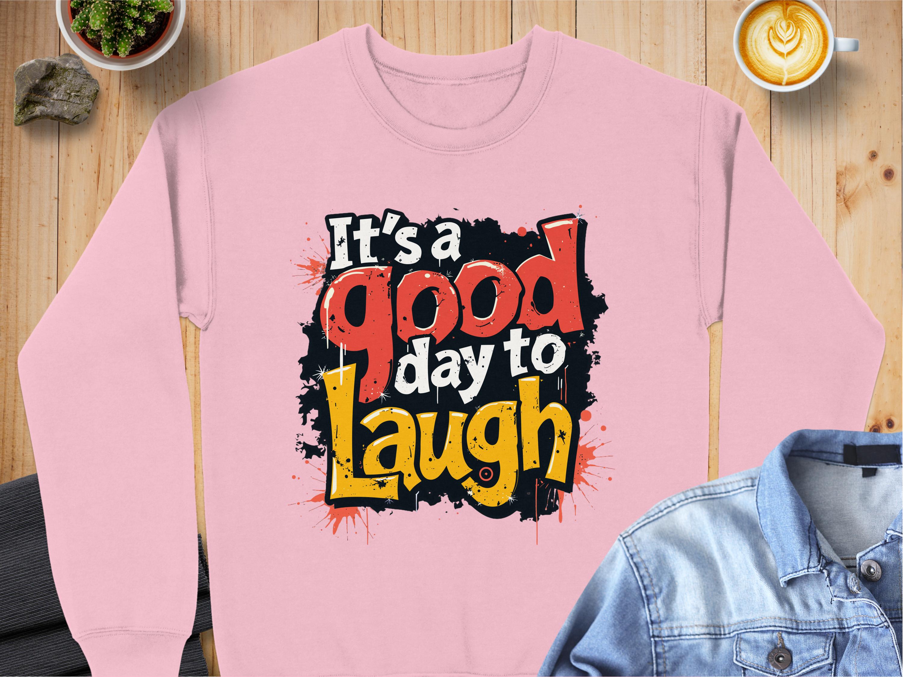 On a wooden surface, Garment Graphics unisex Graphic Art Sweatshirt in pink reads Its a Good Day to Laugh in black, red, and yellow. Its paired with a latte, denim jacket, and two small potted plants.