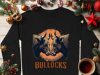 The Garment Graphics black sweatshirt, featuring a bullocks graphic print with two bulls in an orange circle and BULLOCKS boldly below, sits stylishly on a light wooden table surrounded by festive decorations and a steaming cup of coffee.