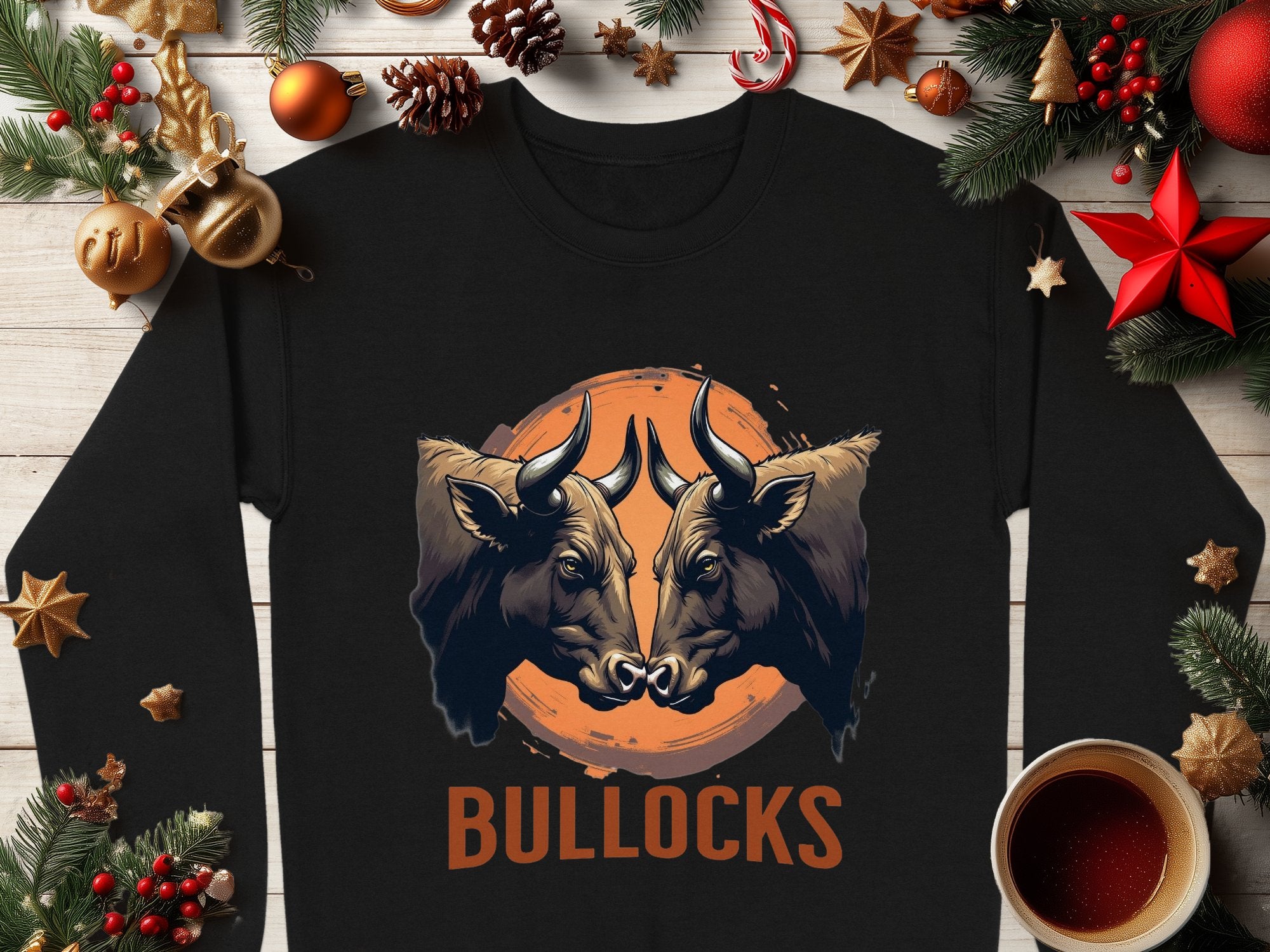 The Garment Graphics black sweatshirt, featuring a bullocks graphic print with two bulls in an orange circle and BULLOCKS boldly below, sits stylishly on a light wooden table surrounded by festive decorations and a steaming cup of coffee.