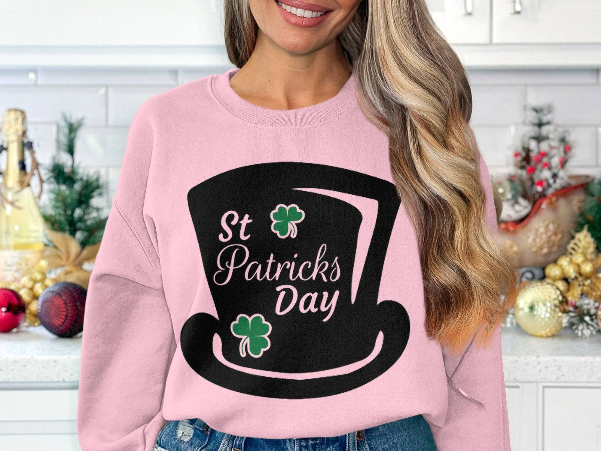 A woman exudes festive vibes in her Garment Graphics pink sweater featuring a St Patricks Day black top hat design with shamrocks, styled with loose wavy hair amidst vibrant kitchen decorations and a countertop.