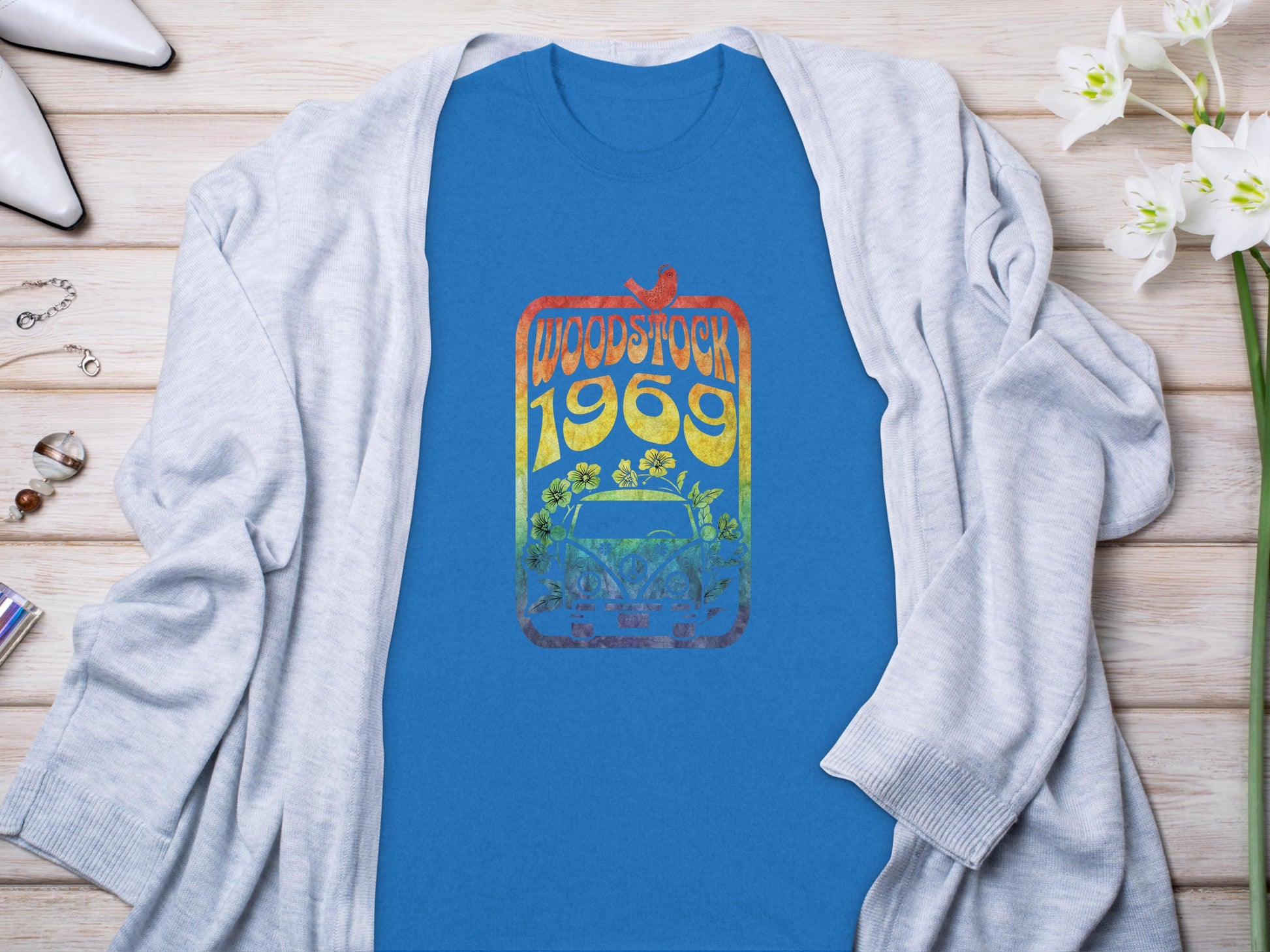 Laid on a wooden surface, the Garment Graphics blue t-shirt with a Woodstock 1969 design showcases a bird, guitar, and flowers. Perfect for music lovers and a retro vibe, its paired with a gray cardigan, white heels, flowers, perfume bottle, and bracelet.