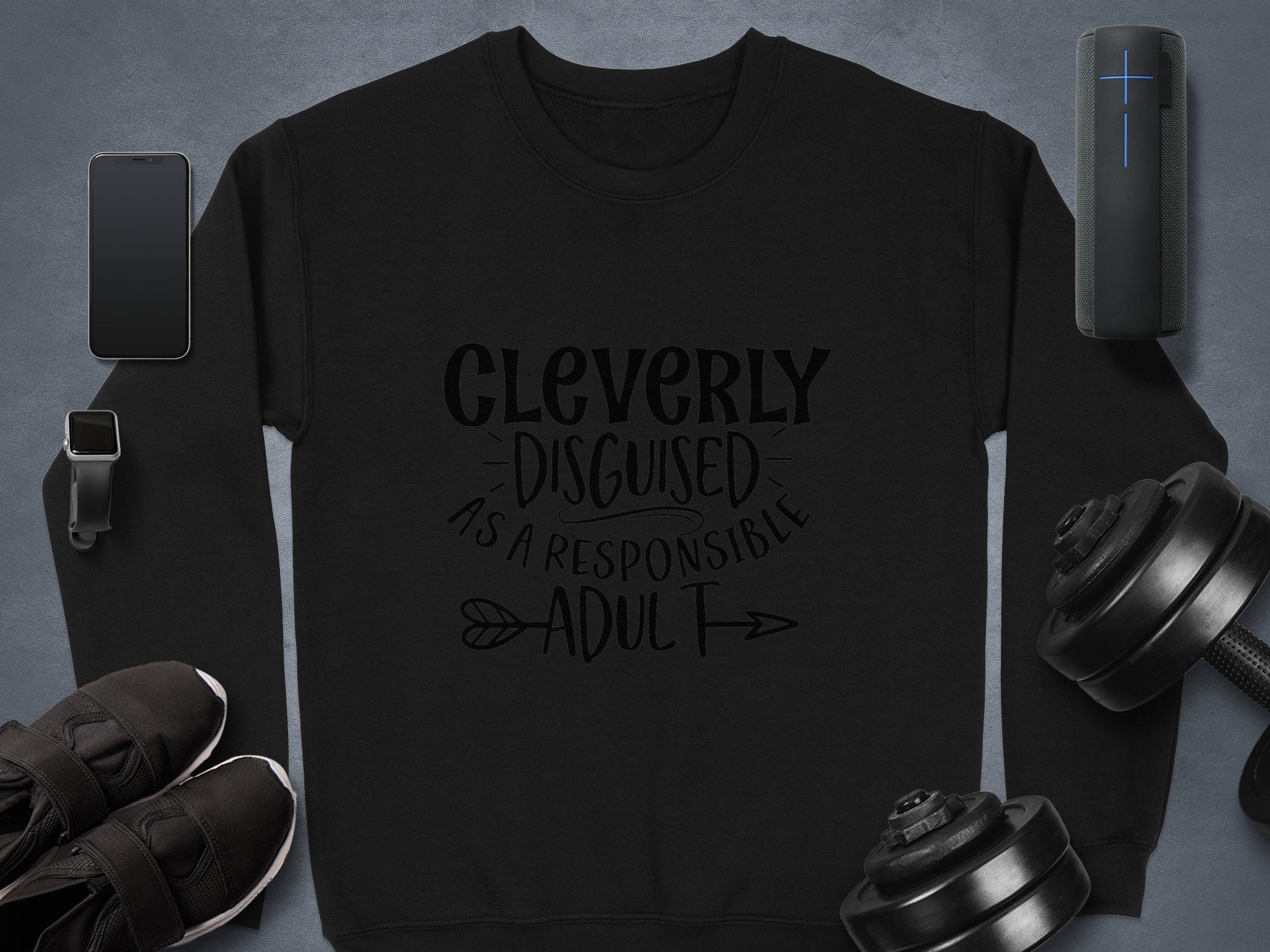 A black Garment Graphics sweatshirt humorously states, Cleverly disguised as a responsible adult in a stylized font. Featured on a gray surface are surrounding items like a smartphone, portable speaker, smartwatch, shoes, and adjustable dumbbells. Perfect for any unisex wardrobe.