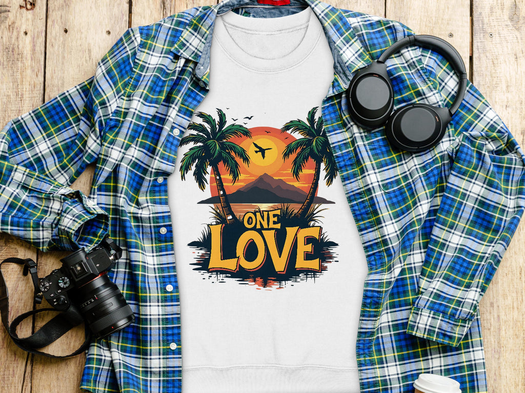 The Garment Graphics white One Love sweatshirt, unisex sizing, features a tropical scene with a sunset between palm trees and a bird. Its paired with a plaid shirt, headphones, camera, and coffee cup on wood.