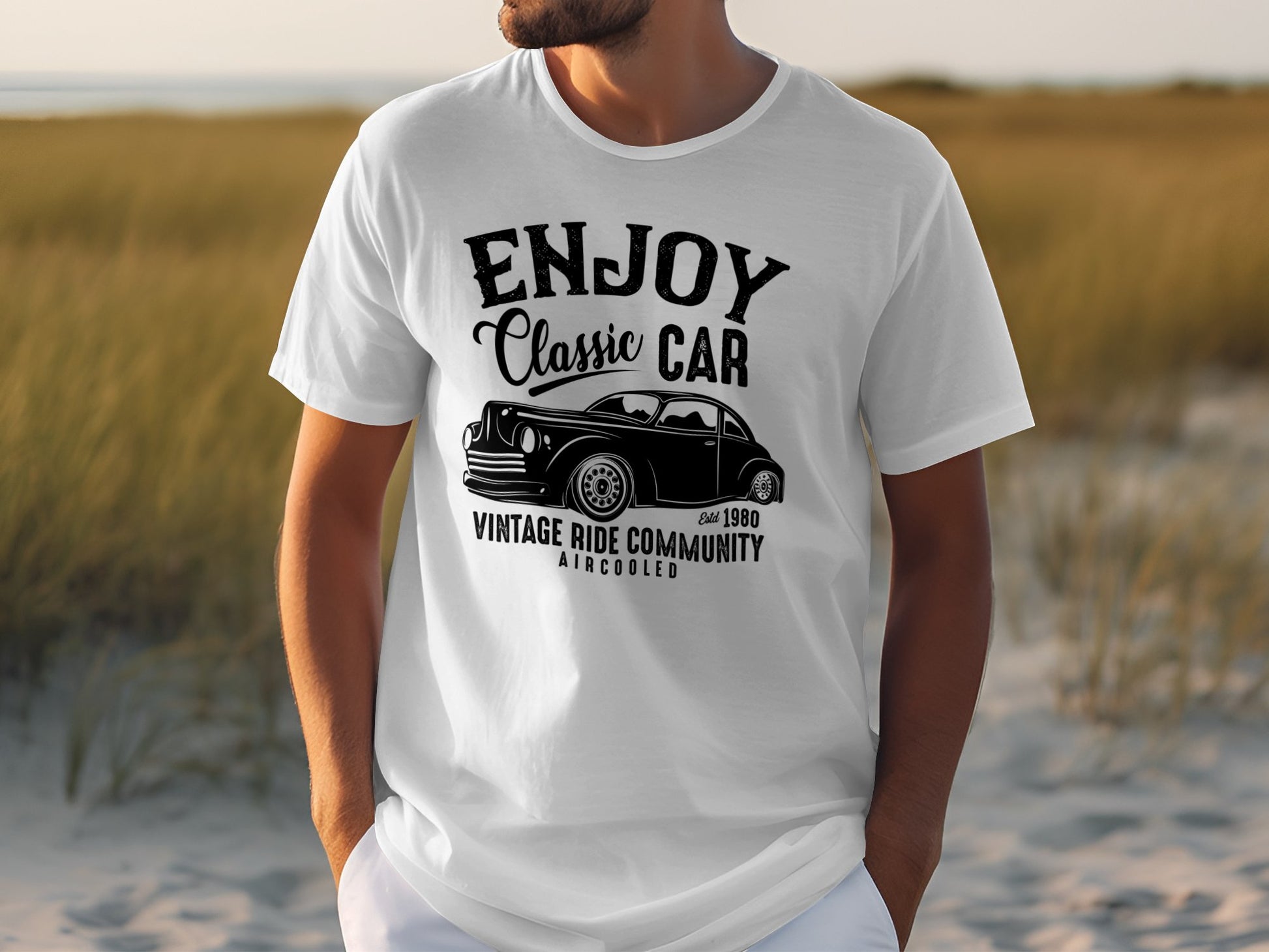 A person wears a white Garment Graphics T-shirt featuring a vintage car illustration and the text ENJOY Classic Car, Vintage Ride Community, Aircooled, estd 1980. Perfect for classic car fans, the scene shows a sandy beach with grass.