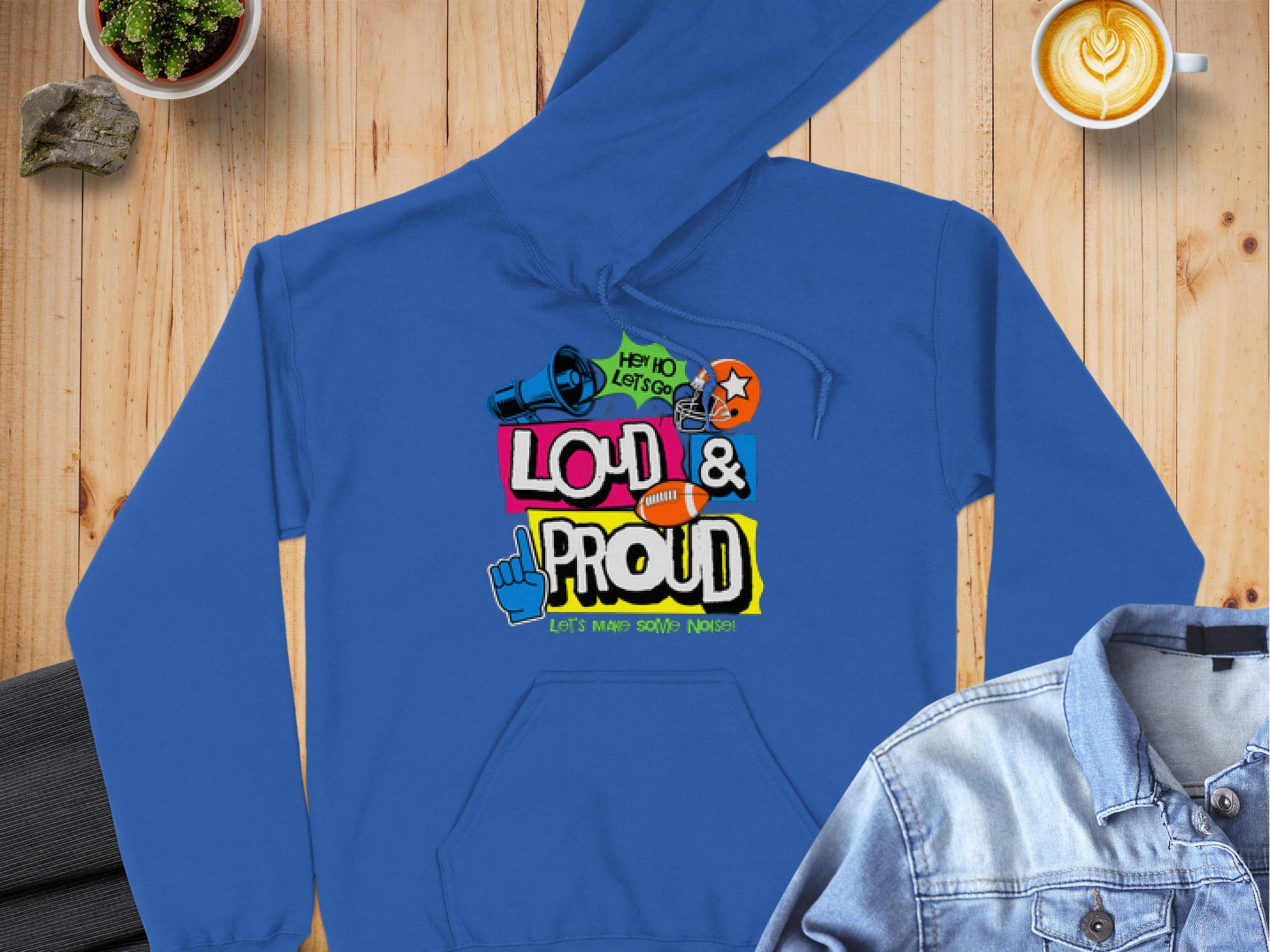 A Garment Graphics blue hoodie with Loud & Proud text, colorful graphics, and speech bubbles rests on wood. Nearby are folded dark pants, a denim jacket, a latte, a plant, and a small rock. This trendy piece from the Lets Make Some Noise Hoodies line makes a bold statement.