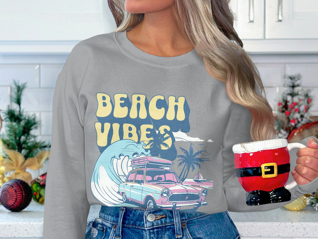 A person is wearing a Garment Graphics white sweatshirt with Beach Vibes in yellow and blue, featuring a pink classic car, surfboards, and palm trees. The retro design shines through the blurred background with greenery, ideal for beach fun.