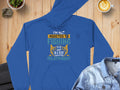 A blue, classic-fit fishing hoodie from Garment Graphics, featuring Im not addicted to fishing; we are just in a very committed relationship, is displayed on a wooden surface with a cup of coffee, denim jacket, and small potted plants nearby.