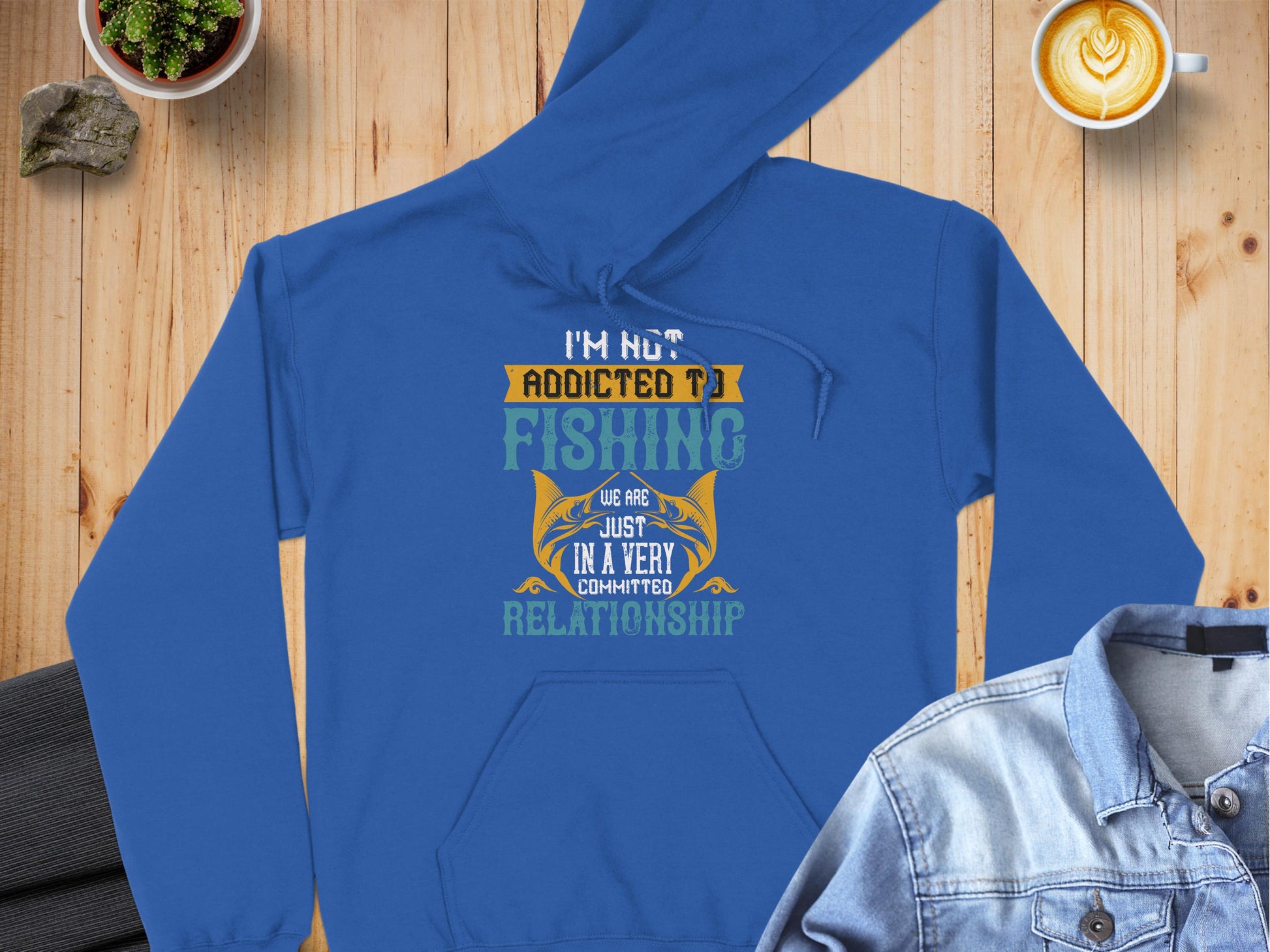 A blue, classic-fit fishing hoodie from Garment Graphics, featuring Im not addicted to fishing; we are just in a very committed relationship, is displayed on a wooden surface with a cup of coffee, denim jacket, and small potted plants nearby.