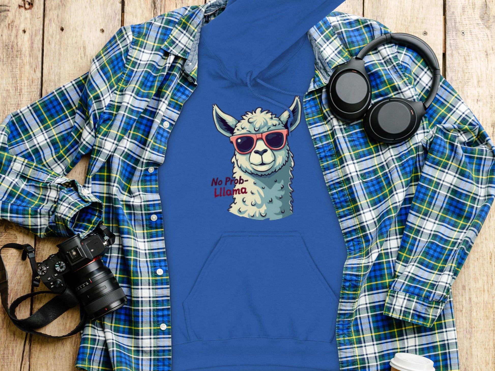 Garment Graphics blue hoodie showcases a sunglasses-wearing llama with the phrase No Prob-Llama. With its classic fit, its styled on wood beside a plaid shirt, headphones, camera, and travel mug.