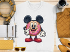 A white sweatshirt from Garment Graphics displays a donut character with mouse-like features in red shoes, holding a pink sprinkled treat. Comes with a plaid scarf, notebook with pen, yellow clutch, sunglasses, mustard jacket, and coffee cup. Unisex sizing offers versatile style.