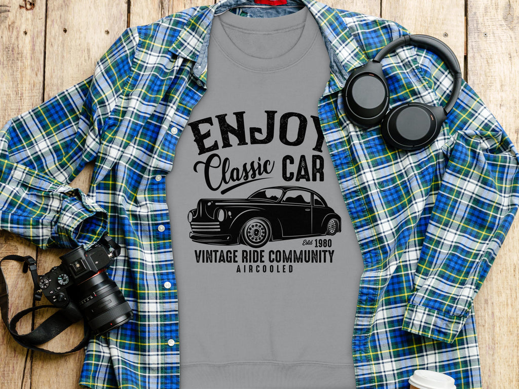 A Garment Graphics gray t-shirt with Enjoy Vintage Ride and a classic car graphic rests on wood, paired with a blue-green plaid shirt. Nearby are headphones, a camera, and a white takeaway cup.