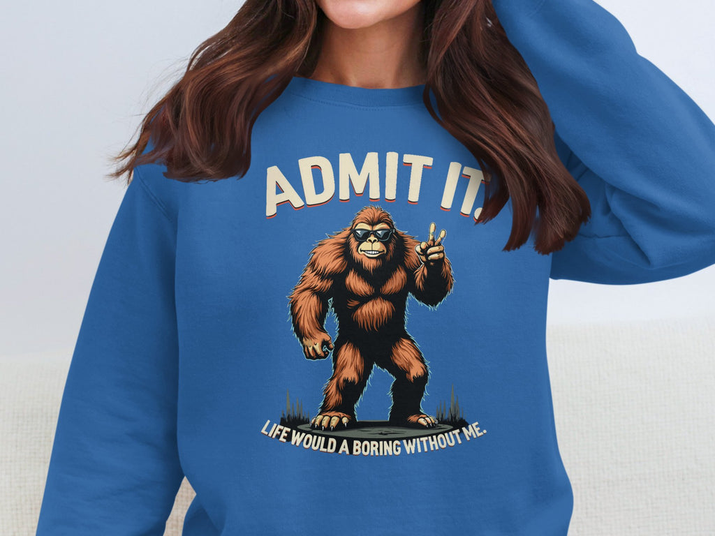 A person wearing a Garment Graphics blue sweatshirt with a playful Bigfoot and ADMIT IT! LIFE WOULD BE BORING WITHOUT ME shows unisex sizing. Their long, dark hair partially covers their hand on their face, enhancing the casual vibe.