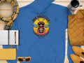 The Garment Graphics Bee Yourself Hoodie in blue, showcasing a bee illustration and Everyone Else Is Taken, Summer Edition text, pairs perfectly with a yellow scarf, black mug, open notebook, yellow clutch, sunglasses, and mustard yellow jacket for a standout look this season!.