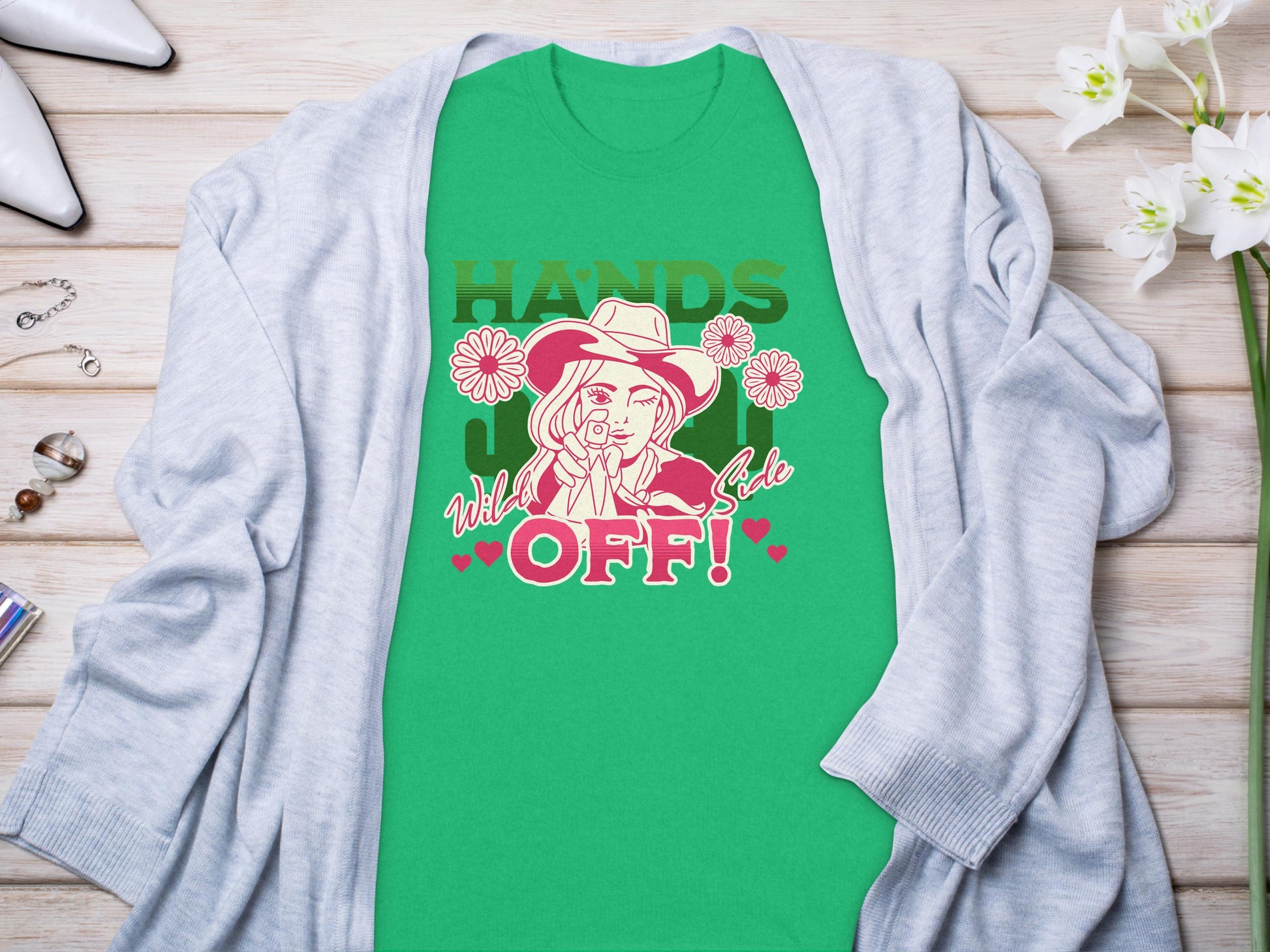 Garment Graphics green t-shirt features a bold design of a person in a hat with HANDS OFF! and Wild & Cute text, surrounded by flowers and hearts. Styled on wood with accessories, it pairs flawlessly with the gray cardigan for a chic classic fit.