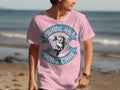 A man on the beach wears a Garment Graphics tee, showcasing a fish and the playful text Fishing Hair Dont Care encircling it. The ocean and sand perfectly highlight this eye-catching design.