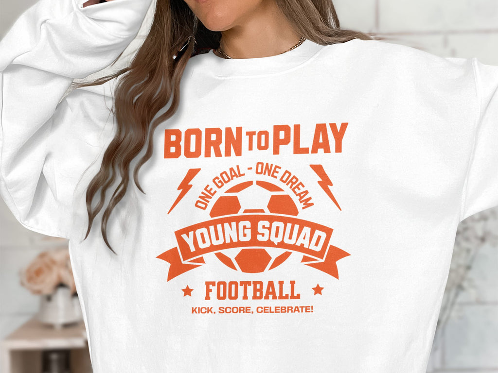 A person wearing a white Garment Graphics football sweatshirt featuring an orange graphic with Born to Play, One Goal - One Dream, Young Squads, Football, Kick, Score, Celebrate! It includes a stylized soccer ball and lightning bolts in unisex sizing.
