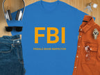 This Garment Graphics blue T-shirt features FBI in bold yellow letters with FEMALE BOOB INSPECTOR below. Its humorously paired with headphones, a keychain, sunglasses, a brown shirt, a wristwatch, and folded jeans on a wooden surface.