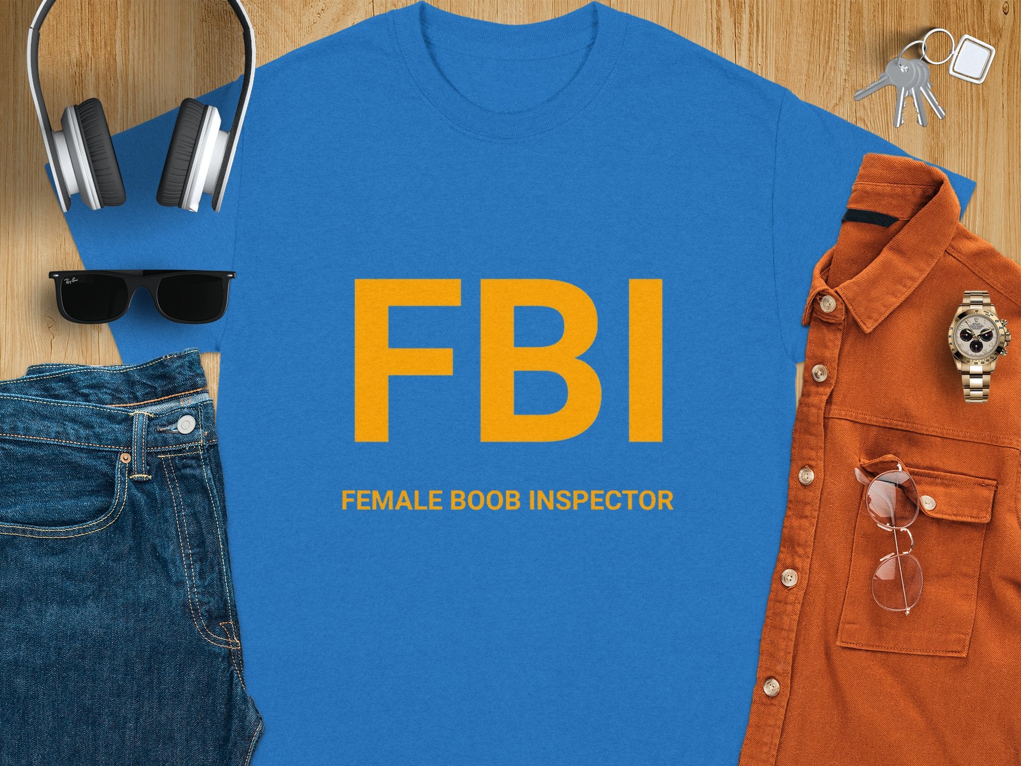 This Garment Graphics blue T-shirt features FBI in bold yellow letters with FEMALE BOOB INSPECTOR below. Its humorously paired with headphones, a keychain, sunglasses, a brown shirt, a wristwatch, and folded jeans on a wooden surface.