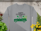 A Garment Graphics gray shirt says Happy St. Patricks Day in black, showcasing a green truck with shamrocks spilling out. Set amid vibrant green and yellow plants on concrete, this festive sweatshirt embodies the holiday spirit.
