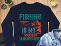 The Garment Graphics navy blue sweatshirt displays Fishing Is My Anger Management in teal and orange, with a fish and fishing lure illustration. Ideal for fishing fans, its styled on a wooden surface with a denim jacket, coffee, and plant enhancing its unique charm.