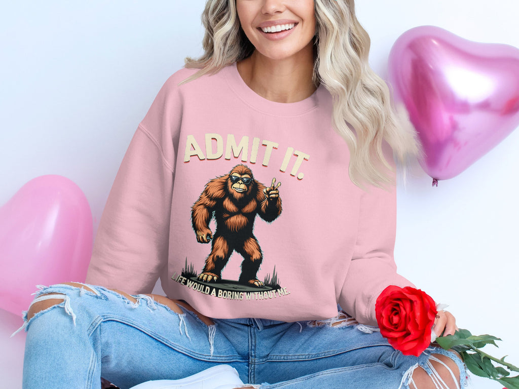 A long-haired blonde woman smiles in casual wear, wearing a pink Garment Graphics sweatshirt featuring a cartoon Bigfoot and the text Admit It. Life Would Be Boring Without Me. She holds a red rose, surrounded by playful pink balloons.