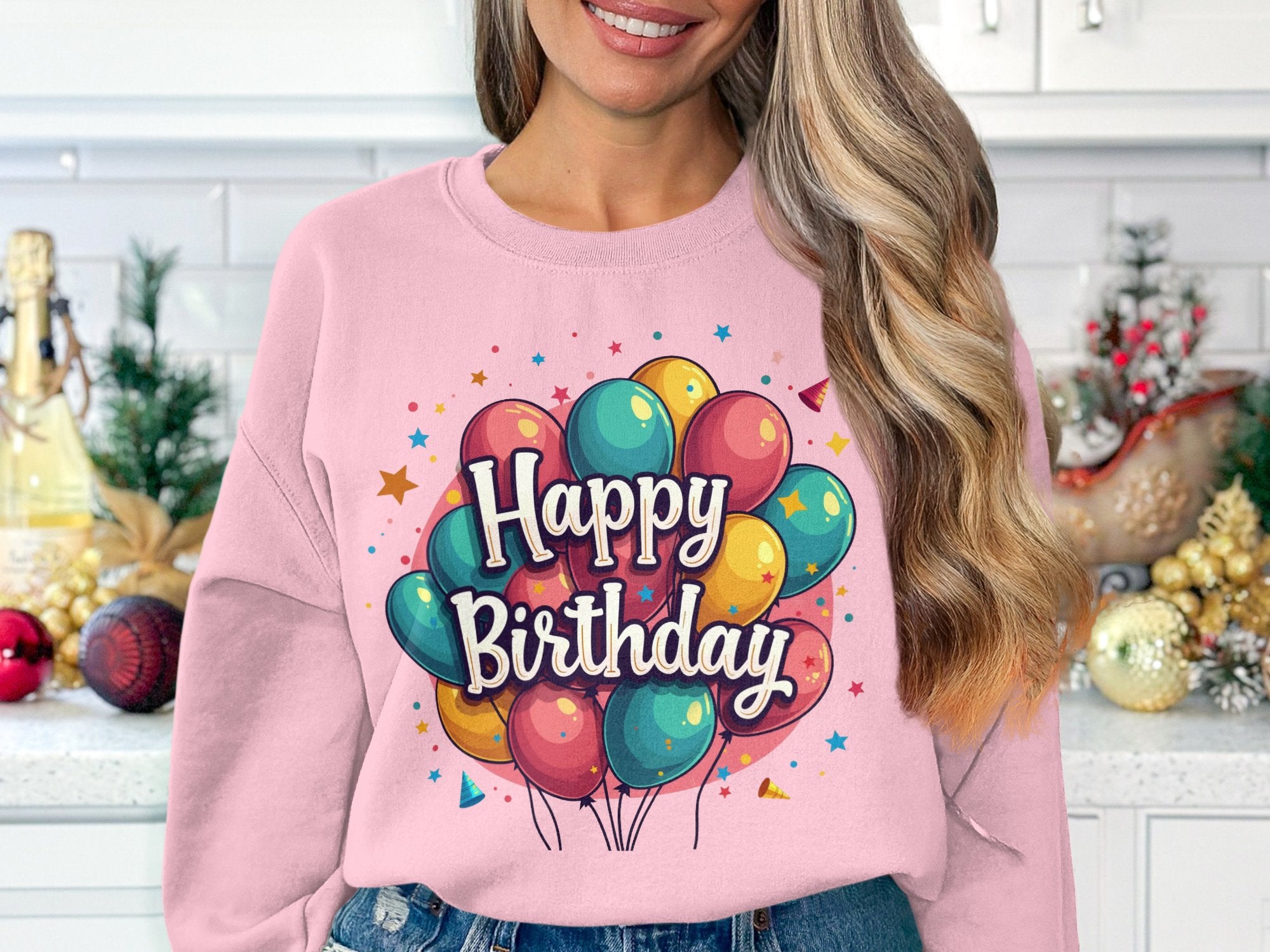 In a kitchen filled with plants and festive ornaments, someone radiates joy wearing Garment Graphics unisex pink sweater. Its colorful Happy Birthday design is adorned with balloons and stars.