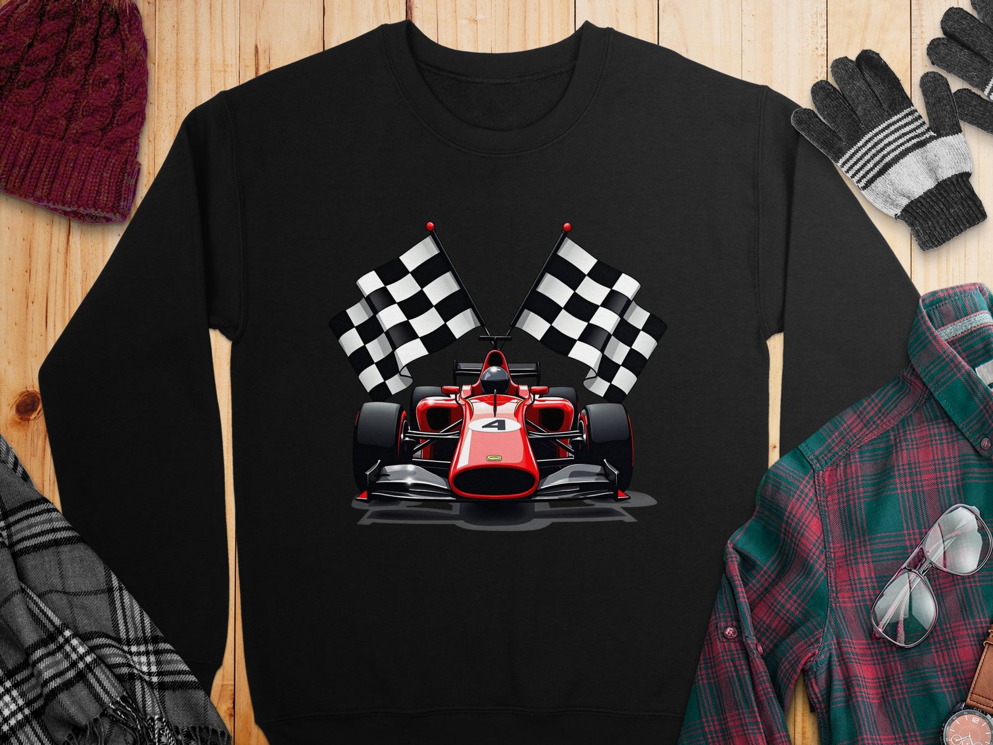 A black Garment Graphics sweatshirt with a red racing car and checkered flags, ideal for motorsport fans. Displayed flat on a wooden surface, its styled with a red knit hat, gloves, plaid shirt, and checked scarf for a lively look.