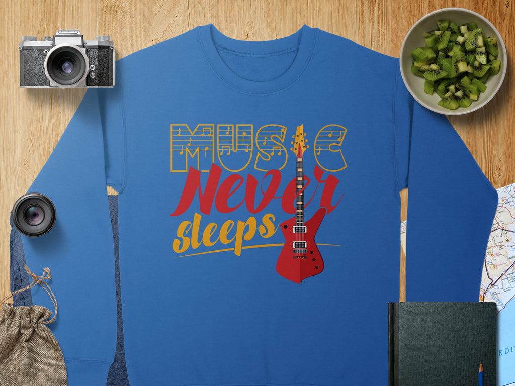 Garment Graphics presents a trendy blue sweatshirt featuring Music Never Sleeps and a vibrant red electric guitar design. Ideal for music lovers, it rests amidst a camera, lens, sack, bowl of sliced kiwi, map, and closed book on a wooden surface.