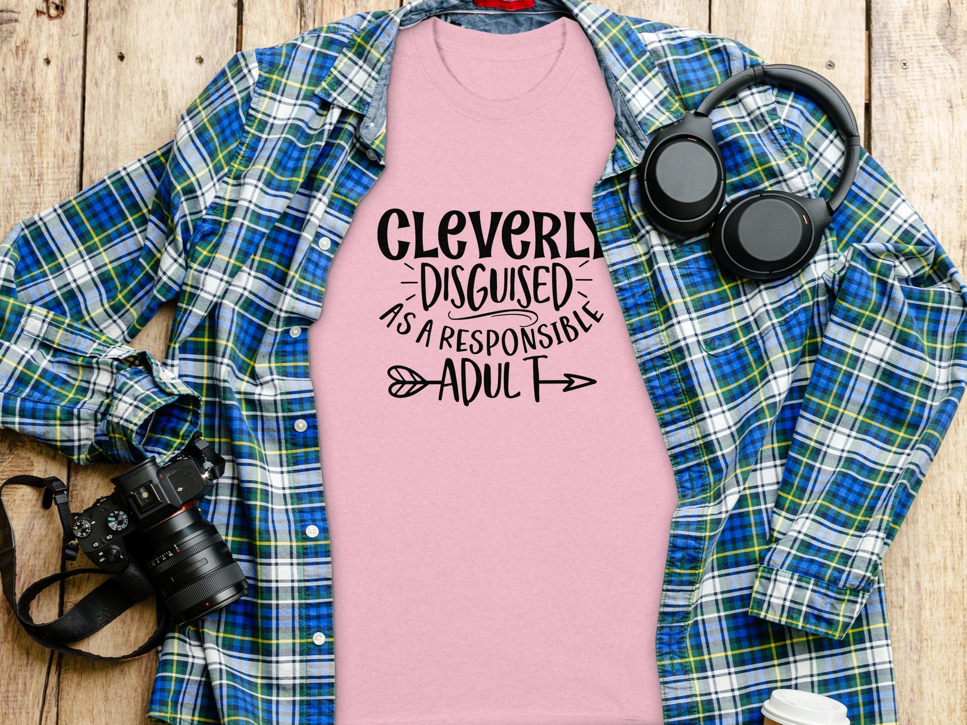 A Garment Graphics pink classic-fit T-shirt with Cleverly Disguised as a Responsible Adult in black text lies on a wooden surface, ideal for casual outings. Its surrounded by a blue and green plaid shirt, black headphones, a lidded coffee cup, and a camera.