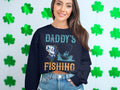 A woman with long dark hair and a broad smile models her favorite Garment Graphics fishing sweatshirt before a shamrock backdrop. The navy blue top features Daddys Fishing Buddy with a fish and boat illustration, capturing the spirit of fishing enthusiasts.