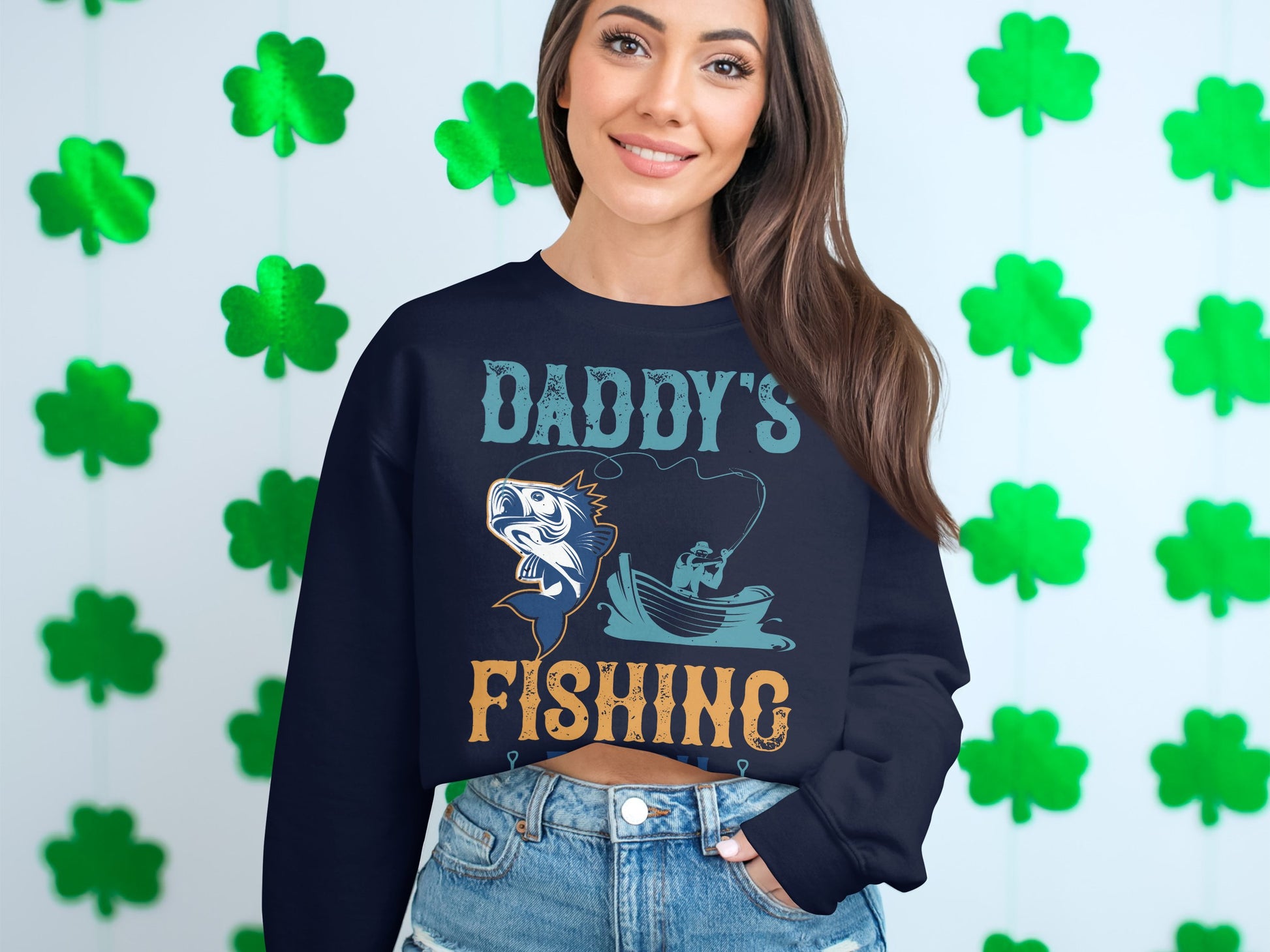 A woman with long dark hair and a broad smile models her favorite Garment Graphics fishing sweatshirt before a shamrock backdrop. The navy blue top features Daddys Fishing Buddy with a fish and boat illustration, capturing the spirit of fishing enthusiasts.