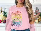 In a kitchen filled with golden ornaments and small Christmas trees, someone exudes retro vibes in a pink Woodstock 1969 sweatshirt by Garment Graphics, featuring a vintage van, flowers, and a bird.