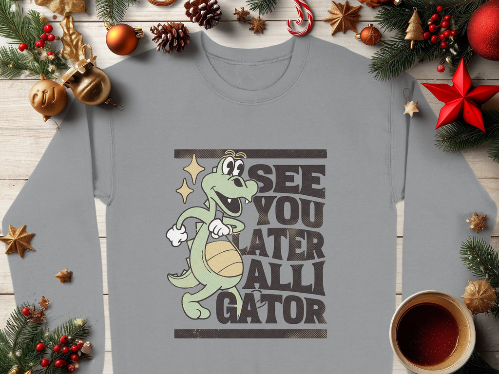 A Garment Graphics gray sweatshirt boasts a playful cartoon gator holding a basketball with See You Later Alligator text. Ideal for graphic sweatshirt fans, it is showcased amid Christmas ornaments, pine branches, and a red star, with coffee adding to the festive vibe.