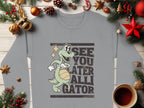 A Garment Graphics gray sweatshirt boasts a playful cartoon gator holding a basketball with See You Later Alligator text. Ideal for graphic sweatshirt fans, it is showcased amid Christmas ornaments, pine branches, and a red star, with coffee adding to the festive vibe.