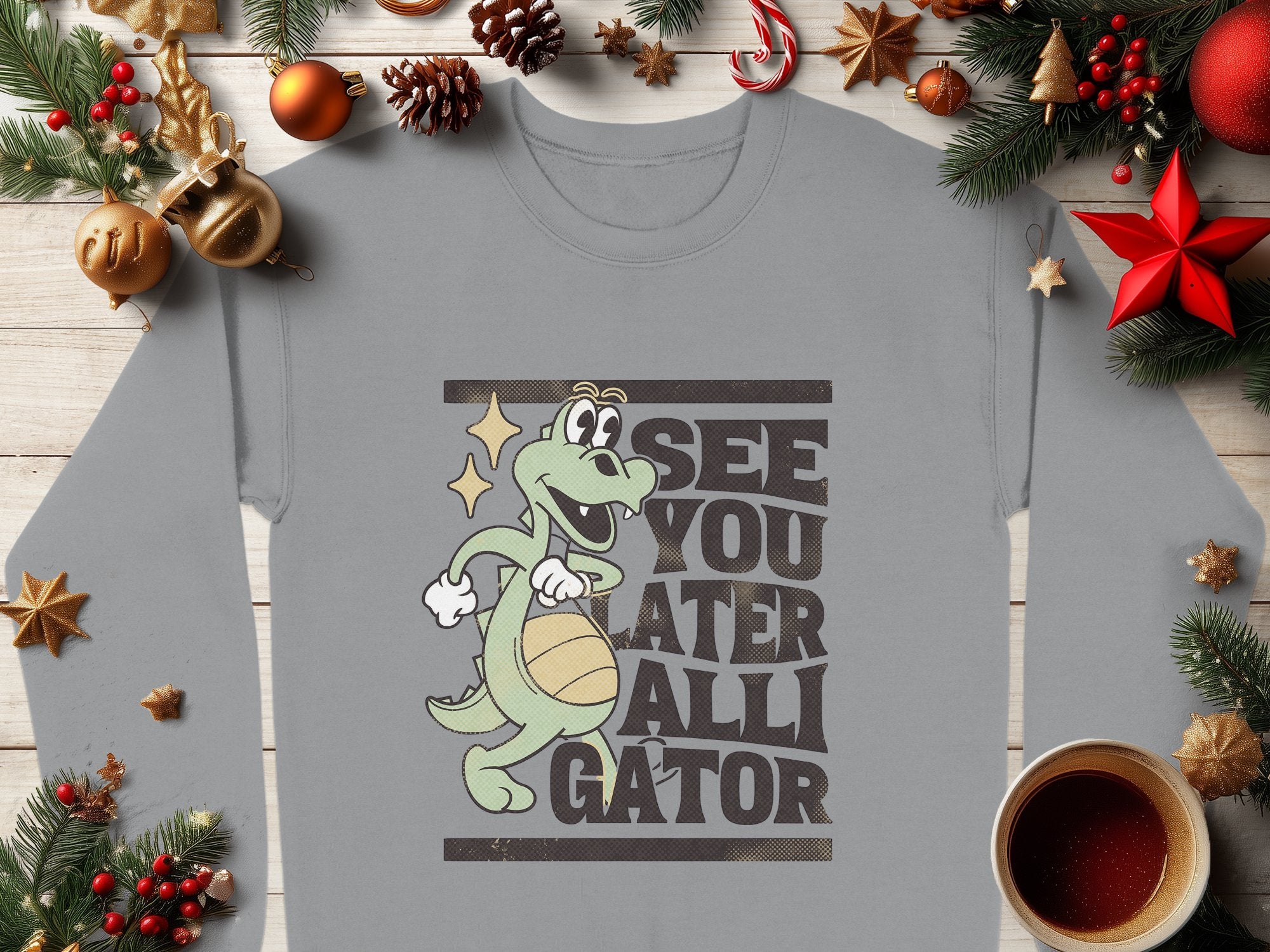 A Garment Graphics gray sweatshirt boasts a playful cartoon gator holding a basketball with See You Later Alligator text. Ideal for graphic sweatshirt fans, it is showcased amid Christmas ornaments, pine branches, and a red star, with coffee adding to the festive vibe.
