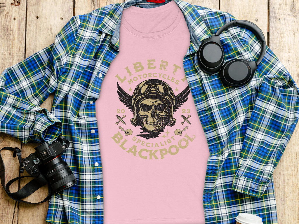 A pink T-shirt from Garment Graphics featuring a skull and wings design with Liberty Motorcycles Specialist Blackpool is displayed on wood. Paired with a blue-green plaid shirt, headphones, camera, and coffee cup—ideal for fans of Blackpool-themed apparel.