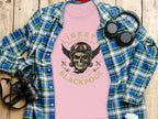 A pink T-shirt from Garment Graphics featuring a skull and wings design with Liberty Motorcycles Specialist Blackpool is displayed on wood. Paired with a blue-green plaid shirt, headphones, camera, and coffee cup—ideal for fans of Blackpool-themed apparel.