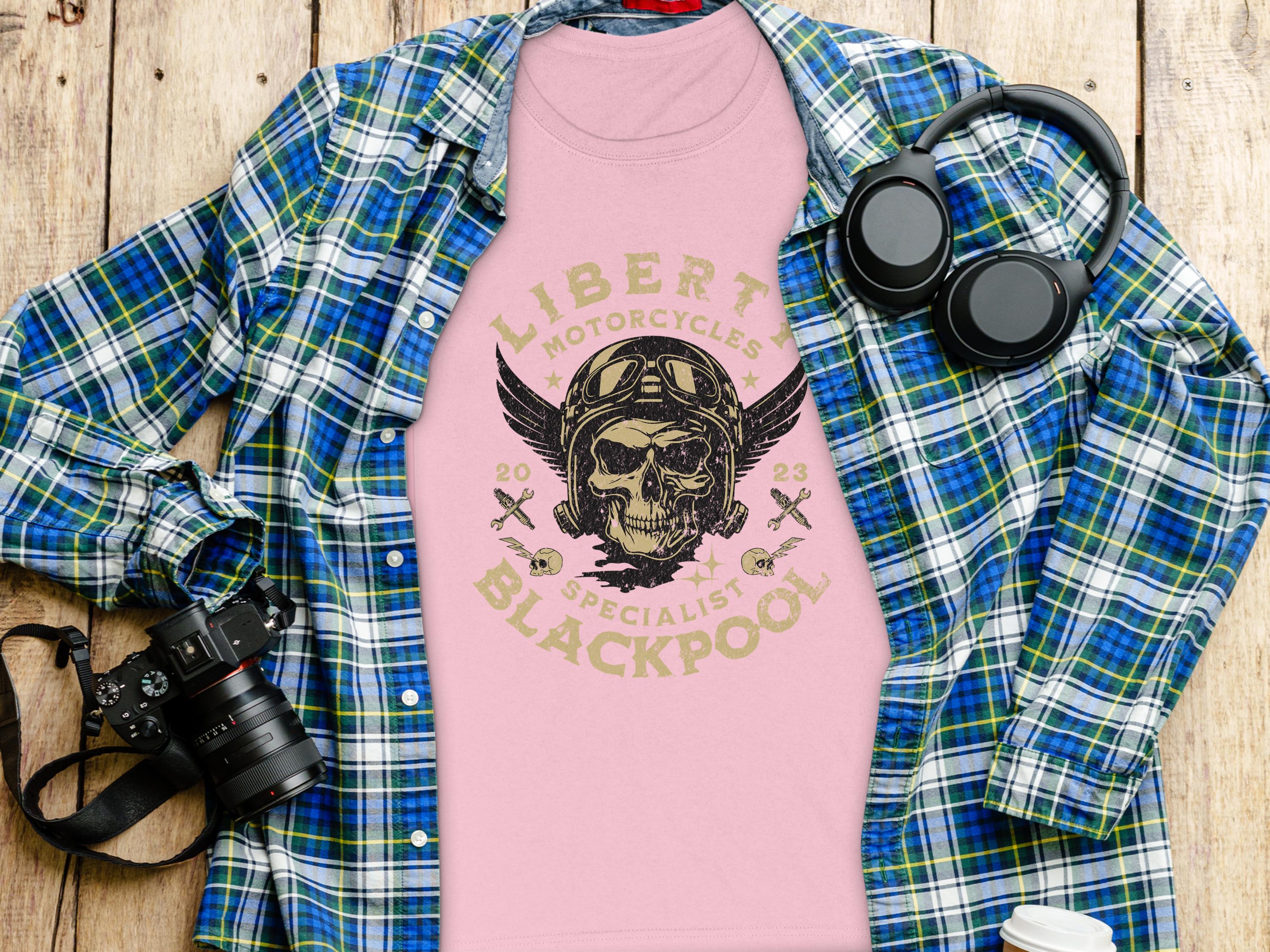 A pink T-shirt from Garment Graphics featuring a skull and wings design with Liberty Motorcycles Specialist Blackpool is displayed on wood. Paired with a blue-green plaid shirt, headphones, camera, and coffee cup—ideal for fans of Blackpool-themed apparel.