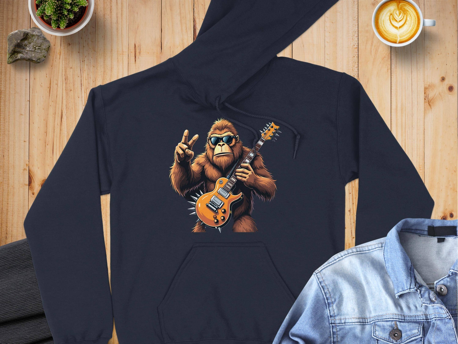 Garment Graphics black electric guitar hoodie features a Bigfoot jam session with a peace sign. Styled on wood beside a denim jacket, cappuccino, plant, and rock, it’s ideal for music lovers craving unique flair.