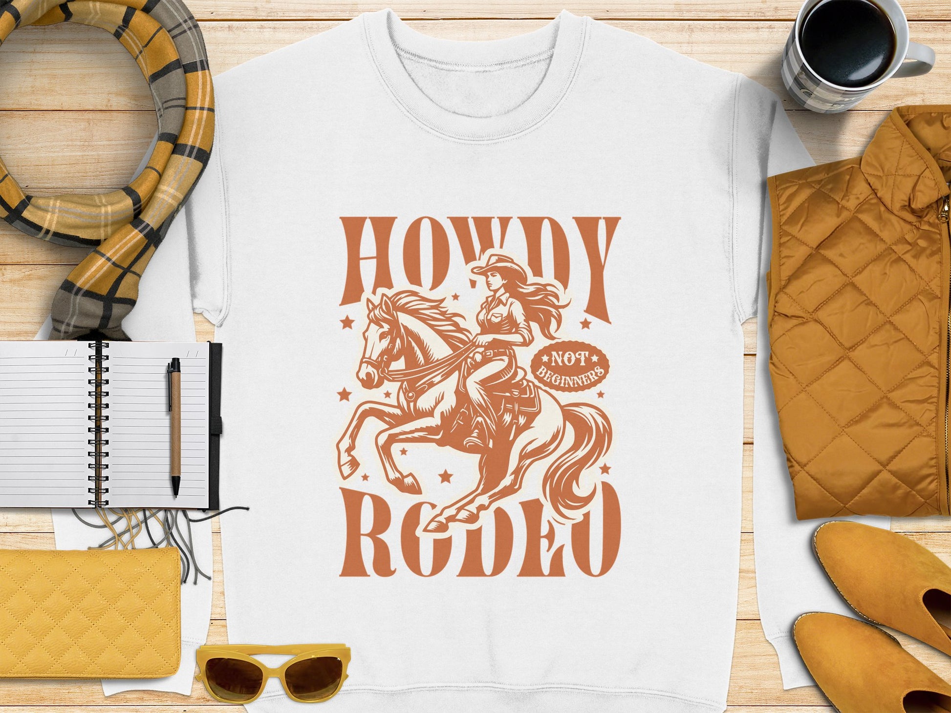 Garment Graphics white t-shirt showcases a cowgirl on horseback and the words Howdy Rodeo, Not For Beginners. Its ideal for rodeo fans embracing their cowboy spirit. The shirt is displayed on wood next to a scarf, notebook, pen, sunglasses, quilted item, and cup.