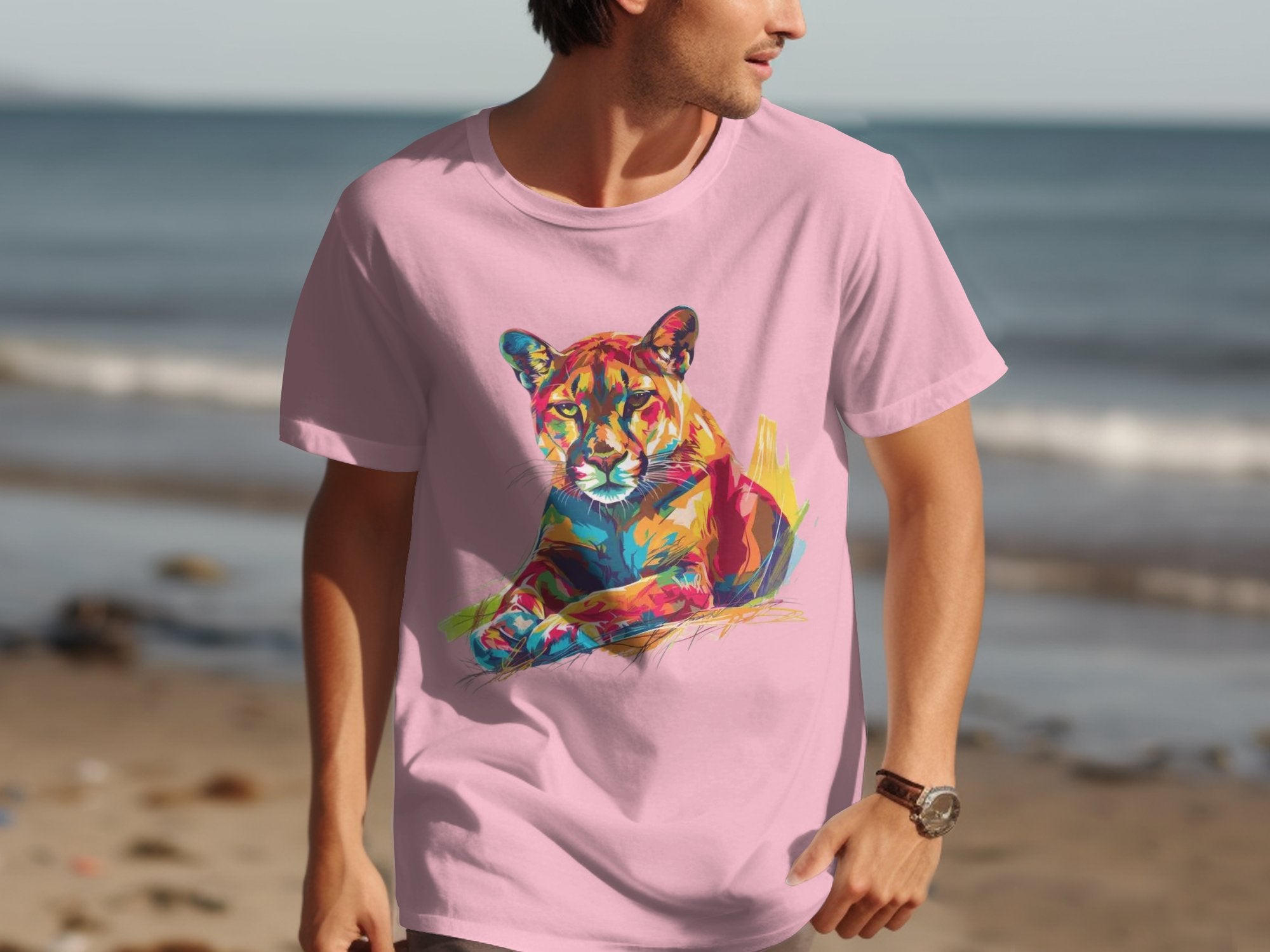 A man on the beach wears a Garment Graphics pink T-shirt with a vibrant lion artwork, making the colorful graphic pop against the sand and sea backdrop.