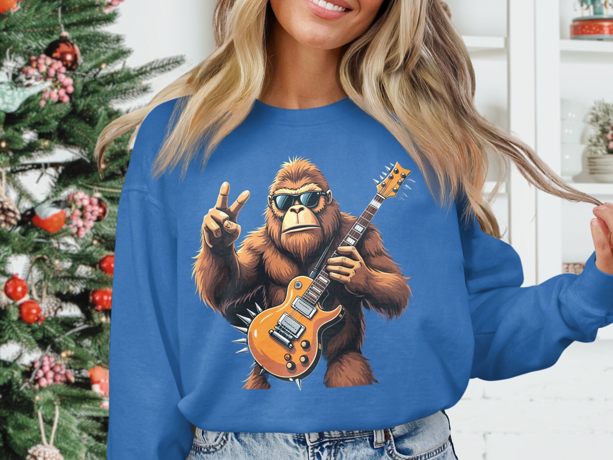 A person wearing a blue Garment Graphics sweatshirt for music lovers, featuring Bigfoot jamming on a guitar with a peace sign and a decorated Christmas tree in the background.