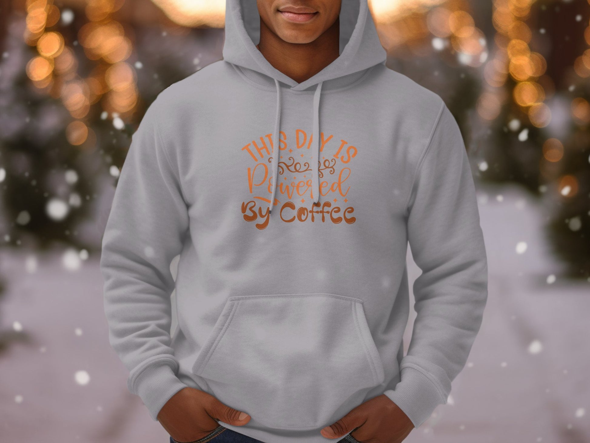 A person wearing a stylish gray Garment Graphics hoodie with This Day Is Powered by Coffee in orange and brown, ideal for coffee lovers. The background of blurred lights and snowy scenery adds to the cozy vibe of this trendy Coffee Hoodie.